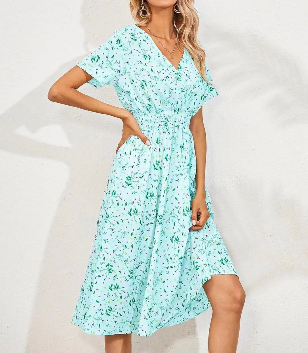 A woman models a light blue V-neck midi dress with a delicate ditsy floral print, short flutter sleeves, and a cinched smocked waist, shown in a three-quarter view against a textured wall.