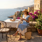 Woman in a light blue Zethri Romantic Lace Appliqué Layered Knit Dress, featuring a deep V-neck and pleated hem, enjoying tea on a sunlit Mediterranean terrace.