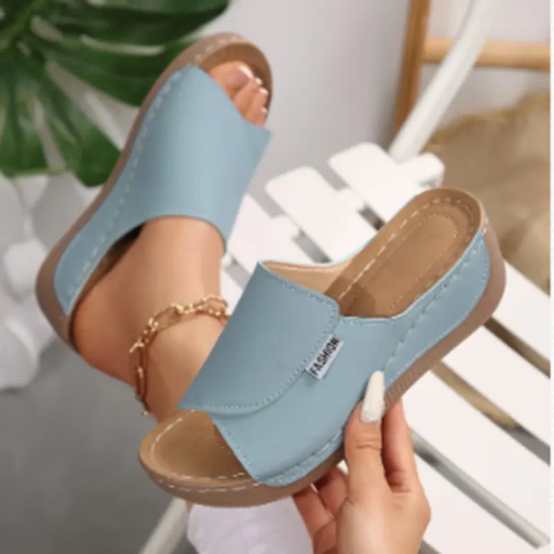 Light blue comfortable platform slide sandals with an open toe and a single wide strap, shown on a foot and being held, featuring a textured wedge sole and contrast stitching.