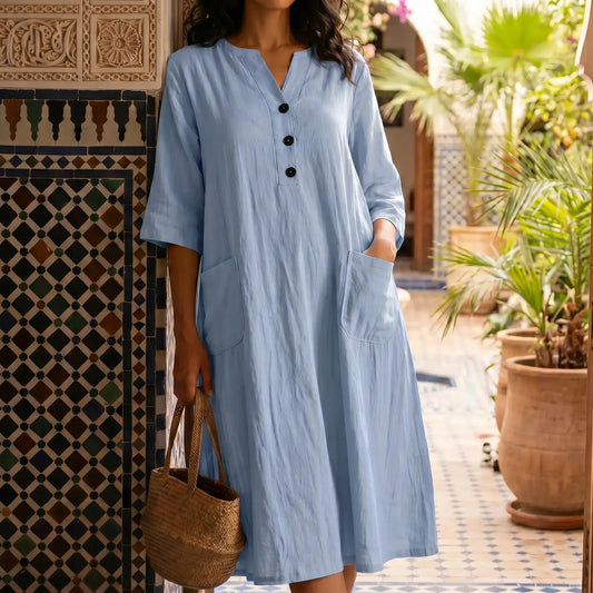 Model wearing a serene light blue V-neck midi dress with half-length sleeves and three dark buttons, showcasing the crinkled texture and front patch pockets, in an elegant outdoor patio setting.