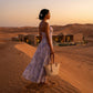Woman wearing the Zevrak lilac floral mesh maxi dress with a ruched bodice and flowing A-line skirt, seen from a three-quarter back view, standing on a desert dune at sunset, holding a woven bag.
