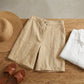 Linara high-waisted linen blend Bermuda shorts in beige, styled flat lay on wood with straw hat and leather sandals.