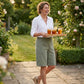 Linara high waisted linen Bermuda shorts in sage green, worn by a smiling woman walking in a sunny garden with iced tea.