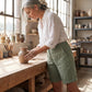 Linara high-waisted linen Bermuda shorts in sage green, worn by a woman shaping clay in a sunlit pottery studio, showing the deep patch pockets and cuffed hem.