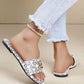 A woman's feet wearing white and black logo print slide sandals with ornate gold buckle, styled with distressed denim jeans and an anklet. The sandal features an all-over repeating monogram pattern.