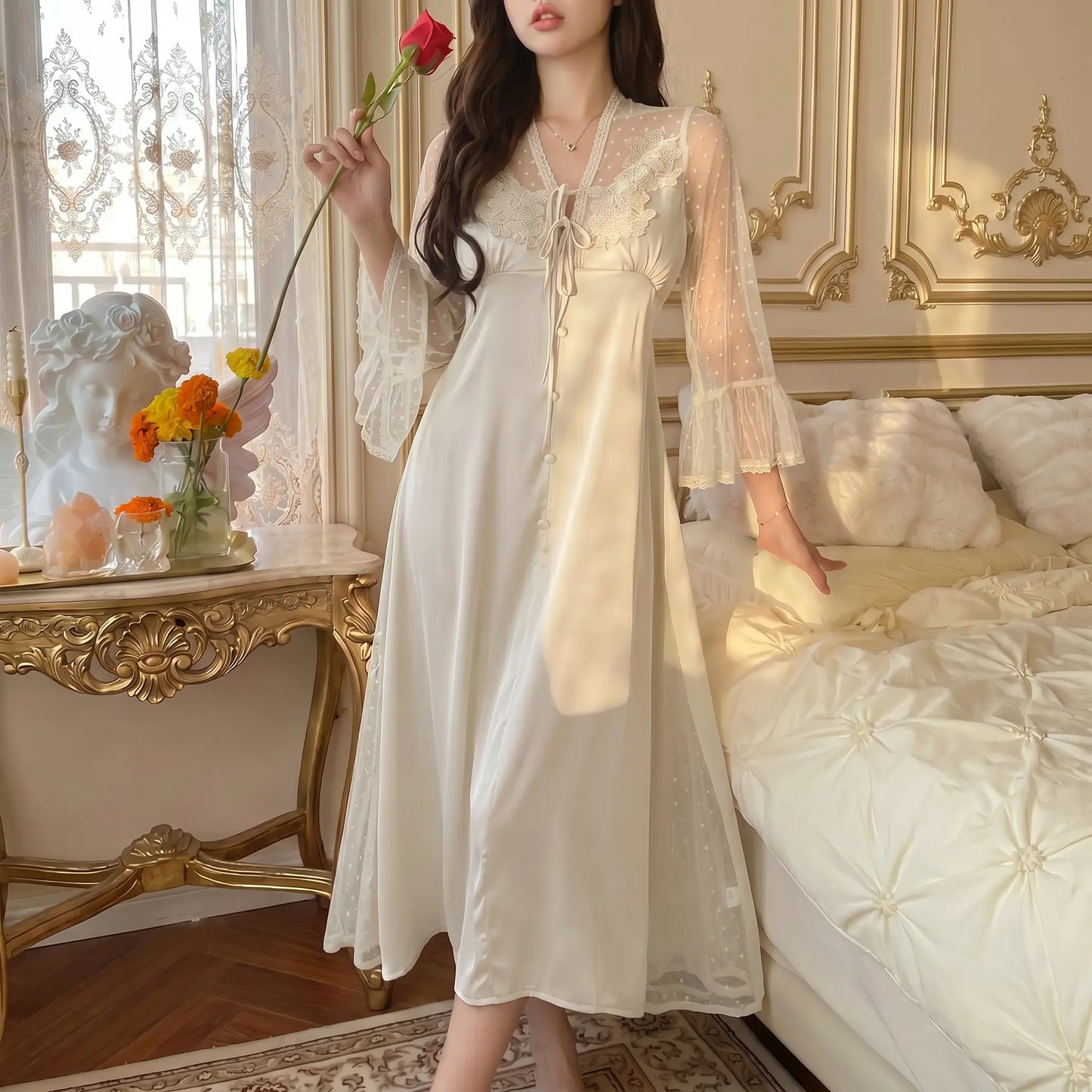 Woman wearing a romantic pearl ivory long satin nightgown with sheer polka-dot bell sleeves, delicate floral lace V-neckline, and a front tie detail, holding a red rose in a sunlit bedroom setting.