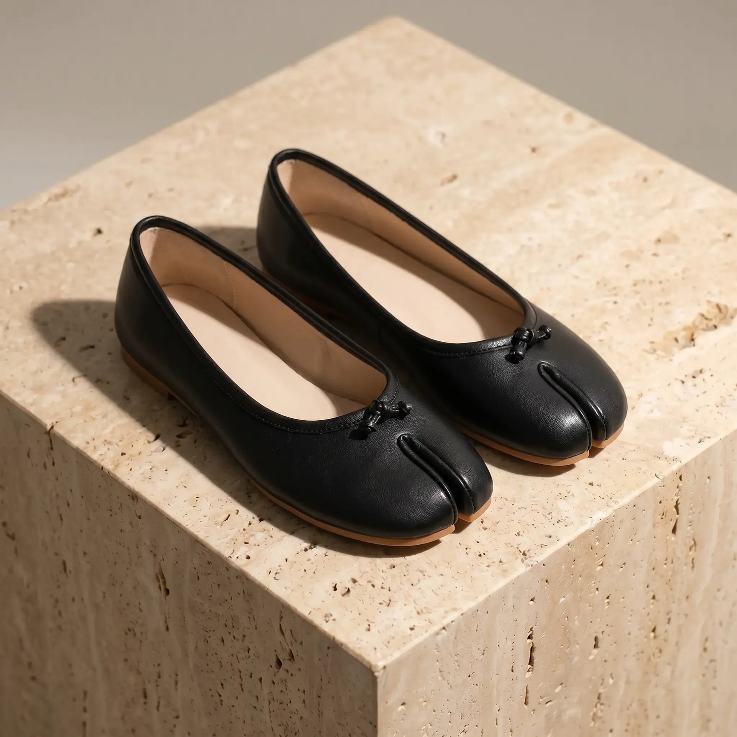 Lurran Midnight Black vegan leather split-toe ballet flats with delicate bows, viewed three-quarter on a textured stone block, showcasing the flat sole.