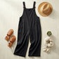 Luvana black cotton linen overalls in a flat-lay style with a straw hat, woven sandals, and a beige scarf on a wooden surface.