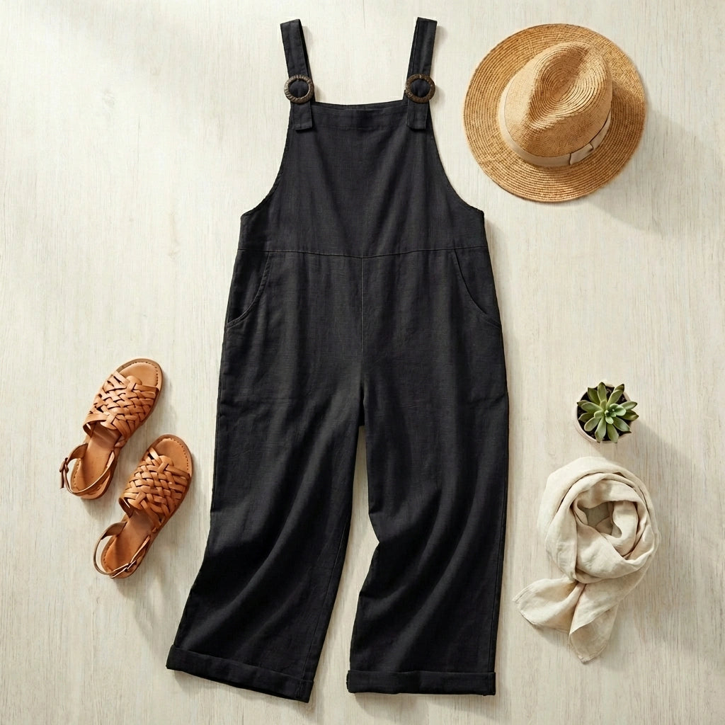 Luvana black cotton linen overalls in a flat-lay style with a straw hat, woven sandals, and a beige scarf on a wooden surface.