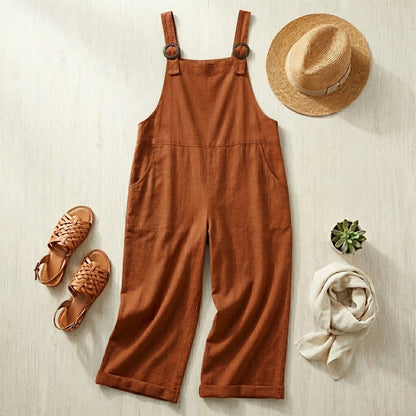 Luvana cotton linen overalls in terracotta red, styled as a flat-lay with a straw hat, sandals, and scarf on a light wood background.
