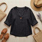 Luvana linen blend blouse in black with V-neck, styled in a flat-lay with a straw hat, woven bag, and sandals on a wooden surface.