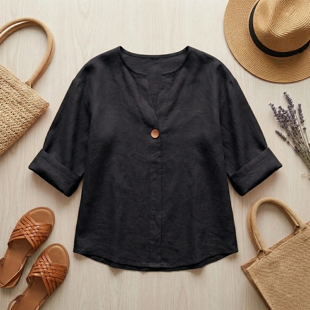 Luvana linen blend blouse in black with V-neck, styled in a flat-lay with a straw hat, woven bag, and sandals on a wooden surface.