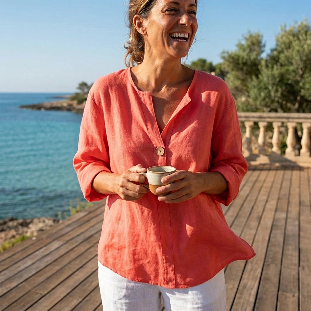 Luvana sunset orange linen blend blouse with V-neck and single-button detail worn by a woman on a sunny deck overlooking the sea.