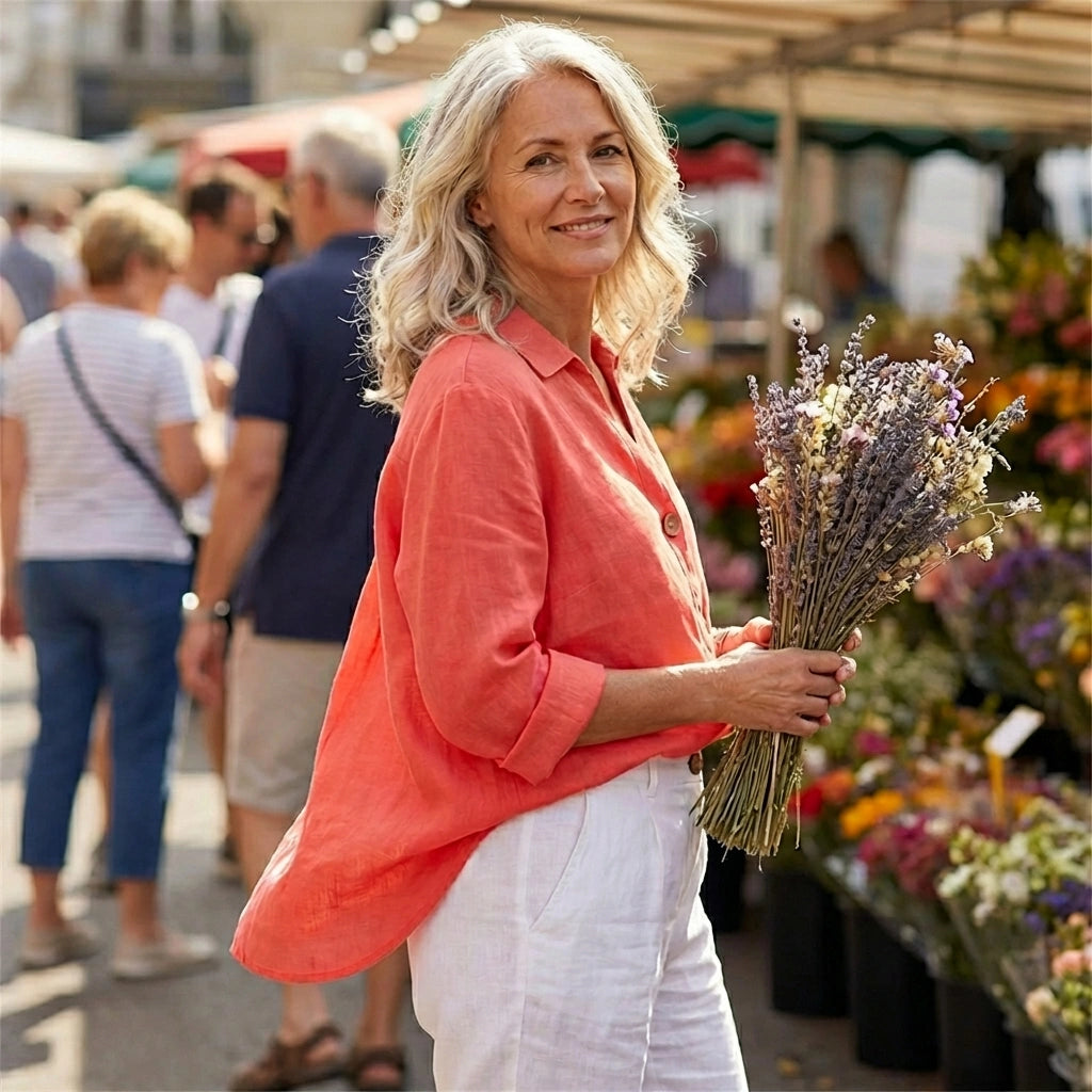 Woman wearing a sunset orange Luvana linen blend blouse with a relaxed V-neck, holding lavender at an outdoor market.
