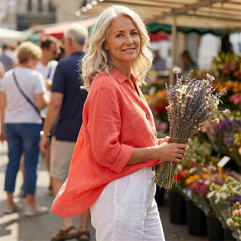 Woman wearing a sunset orange Luvana linen blend blouse with a relaxed V-neck, holding lavender at an outdoor market.