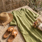 Luvara sage green linen-look V-neck dress with side pockets, styled with a straw hat and leather sandals on a rustic stone surface.