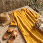 Luvara linen-look v-neck dress in golden ochre, displayed flat with a straw hat and dried flowers on a rustic stone surface.