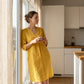Woman wearing a golden ochre Luvara linen-look v-neck dress with 3/4 sleeves, standing in a bright kitchen.