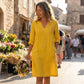 Luvara linen-look V-neck dress in golden ochre with 3/4 sleeves, worn by a woman walking outdoors with a bouquet of flowers.
