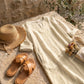 Luvara linen-look V-neck dress in beige styled with a straw hat and leather sandals on a stone surface.
