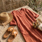Luvara linen-look V-neck dress in burnt terracotta, styled as a flat-lay with a straw hat and leather sandals on a stone surface.