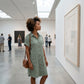 Woman wearing a sage green Luvara linen shirt dress with tortoiseshell buttons and pockets while viewing art in a modern gallery.