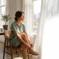 Woman wearing a Luvara sage green linen shirt dress with tortoiseshell buttons, sitting by a window in a relaxed lifestyle setting.