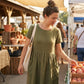 Luvia olive green cotton linen midi dress with pockets worn by a woman at an outdoor market, showing the relaxed fit and natural texture.