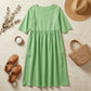 Luvia sage green cotton linen midi dress in a flat-lay lifestyle setting with a straw hat, woven bag, and sandals on a stone surface.
