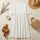 Luvia white cotton linen midi dress in a flat-lay arrangement with a straw hat, sandals, and eucalyptus on a textured stone background.