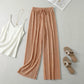 Luvia wide leg pleated plisse trousers in apricot color, styled in a flat-lay with a white camisole, gold necklace, and beige sandals.