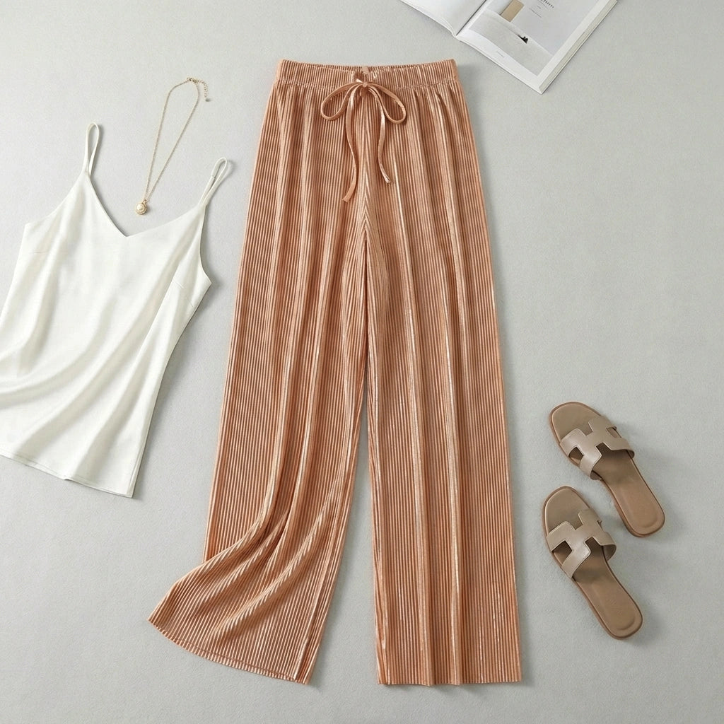Luvia wide leg pleated plisse trousers in apricot color, styled in a flat-lay with a white camisole, gold necklace, and beige sandals.