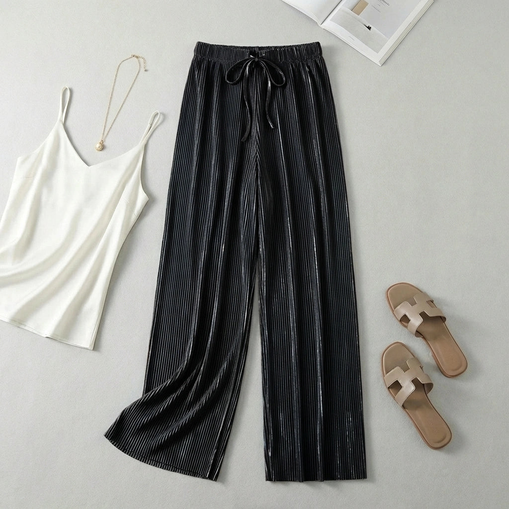 Luvia black wide-leg pleated plissé trousers styled in a flat-lay with a white camisole and beige slide sandals.