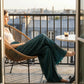 Luvia wide-leg pleated plisse trousers in deep peacock blue, worn by a woman sitting on a balcony with a city view.