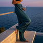 Luvia wide-leg pleated plissé trousers in deep teal with high-waist fit, worn by a woman overlooking the ocean at sunset.