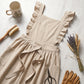 Luvina ruffled cotton pinafore apron in beige with cross-back design, displayed in a cottagecore lifestyle setting with gardening tools.
