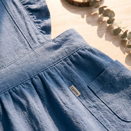 Close-up of Luvina ruffled cotton pinafore apron in blue, showing the side pocket, brand label, and gathered skirt detail.