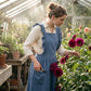 Luvina blue ruffled cotton pinafore apron worn by a woman pruning flowers in a sunlit greenhouse, showing the cross-back design and pockets.