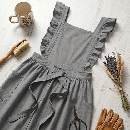 Luvina ruffled cotton pinafore apron in slate gray, displayed in a flat-lay with gardening tools, dried flowers, and a ceramic mug.