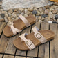 Pair of elegant beige Lyraea cork footbed slide sandals with adjustable antique brass buckle straps, displayed on a rustic wooden surface with pebbles and lace.