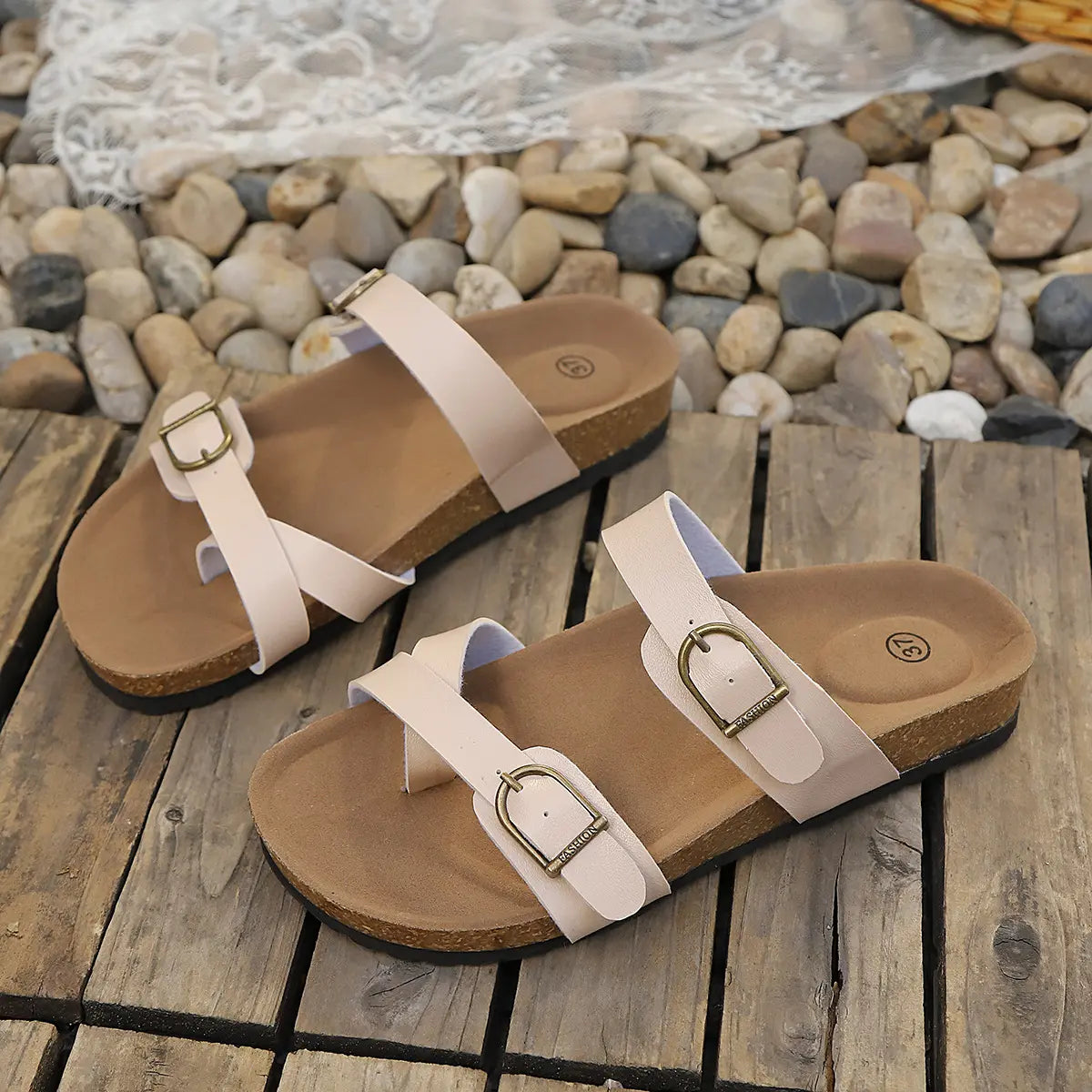 Pair of elegant beige Lyraea cork footbed slide sandals with adjustable antique brass buckle straps, displayed on a rustic wooden surface with pebbles and lace.