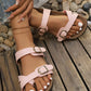 Elegant pink cork footbed slide sandals with dual antique brass buckle straps, shown on a woman's feet with white pedicure, resting on a rustic wooden surface with scattered stones.