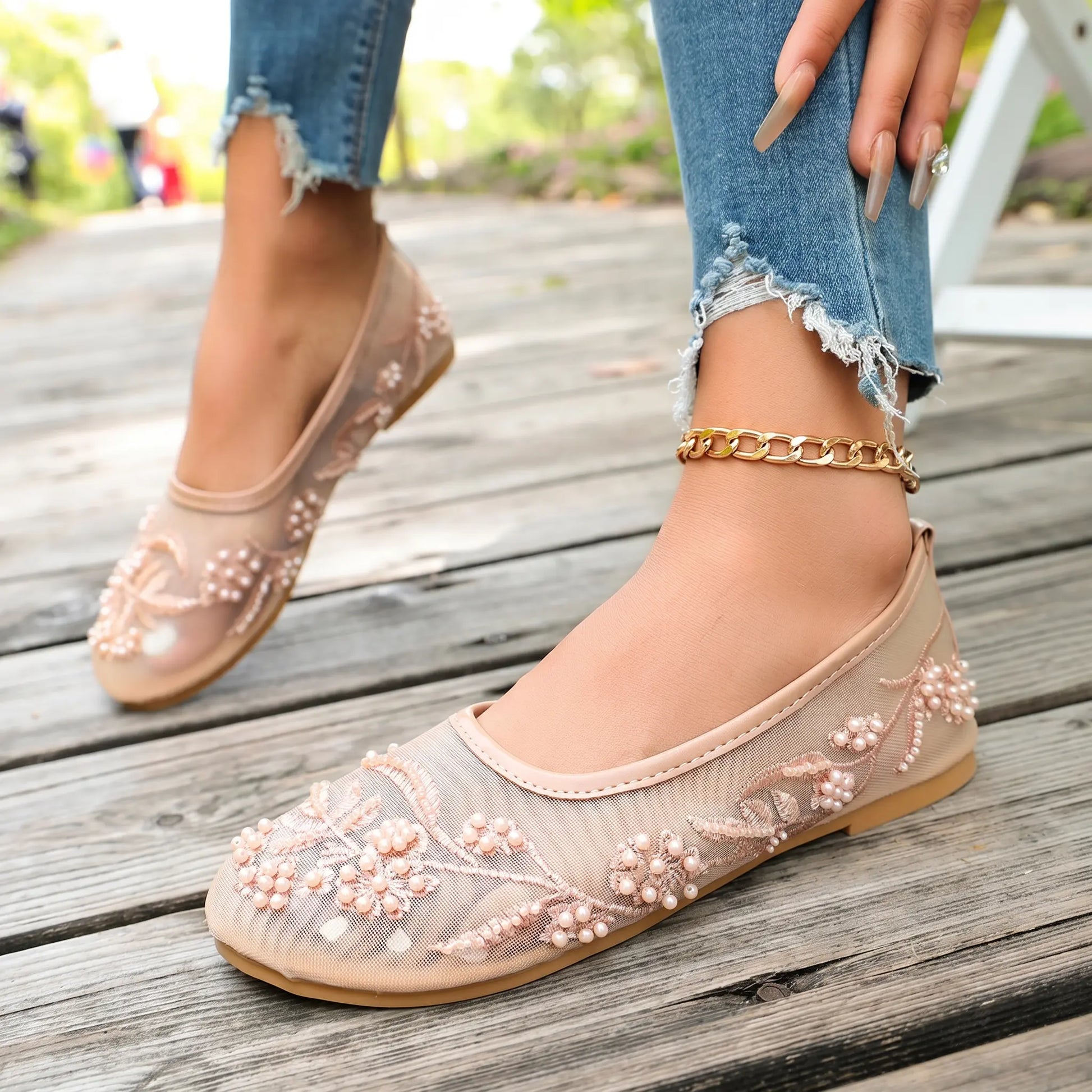 Nude sheer mesh ballet flats adorned with delicate floral embroidery featuring small faux pearls and beads, shown on feet with distressed jeans and an ankle chain. These romantic shoes rest on a wooden walkway.