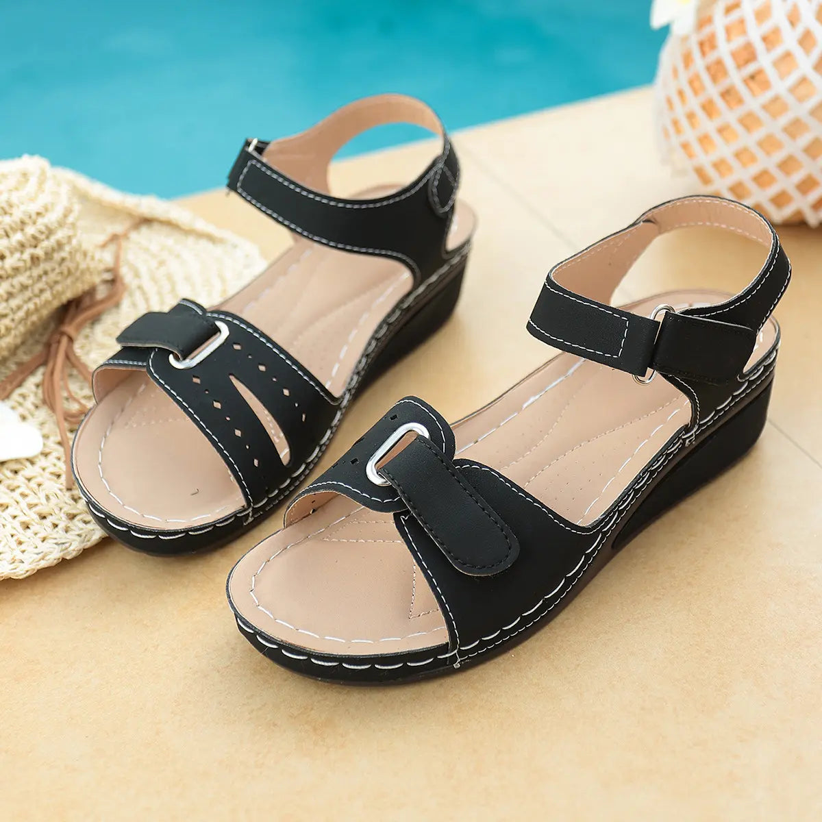 Black Lyraia women's comfort wedge sandals featuring a peep-toe design, adjustable hook-and-loop straps with white contrast stitching, and perforated detailing, shown in a relaxed summer poolside setting.