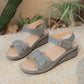 Lyraia women's taupe grey comfort wedge sandals featuring peep-toe, adjustable hook-and-loop straps, and decorative perforated front detail, set on a textured sandy surface with greenery in the background.