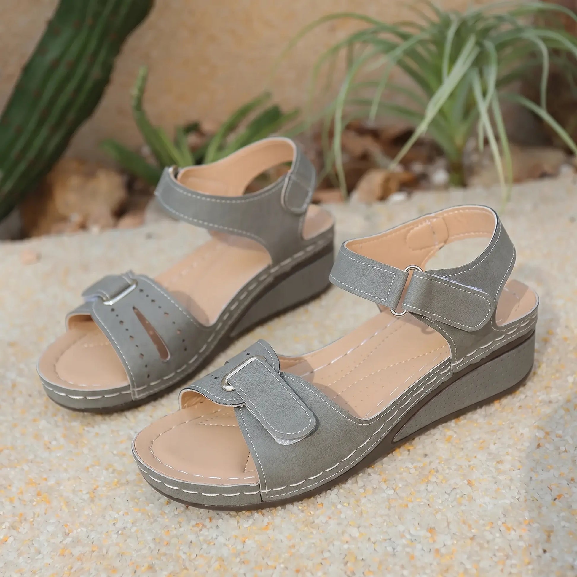 Lyraia women's taupe grey comfort wedge sandals featuring peep-toe, adjustable hook-and-loop straps, and decorative perforated front detail, set on a textured sandy surface with greenery in the background.