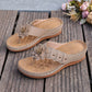 Elegant beige thong sandals featuring a decorative layered fabric flower embellishment, gold studs, and white contrast stitching on a low wedge sole. A pair of Lyraka floral sandals displayed on wooden planks.