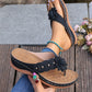 Black Lyraka thong sandals with a black fabric flower embellishment, gold studs, and white contrast stitching, shown on a foot with a turquoise anklet and held in hand, set against a natural outdoor background.