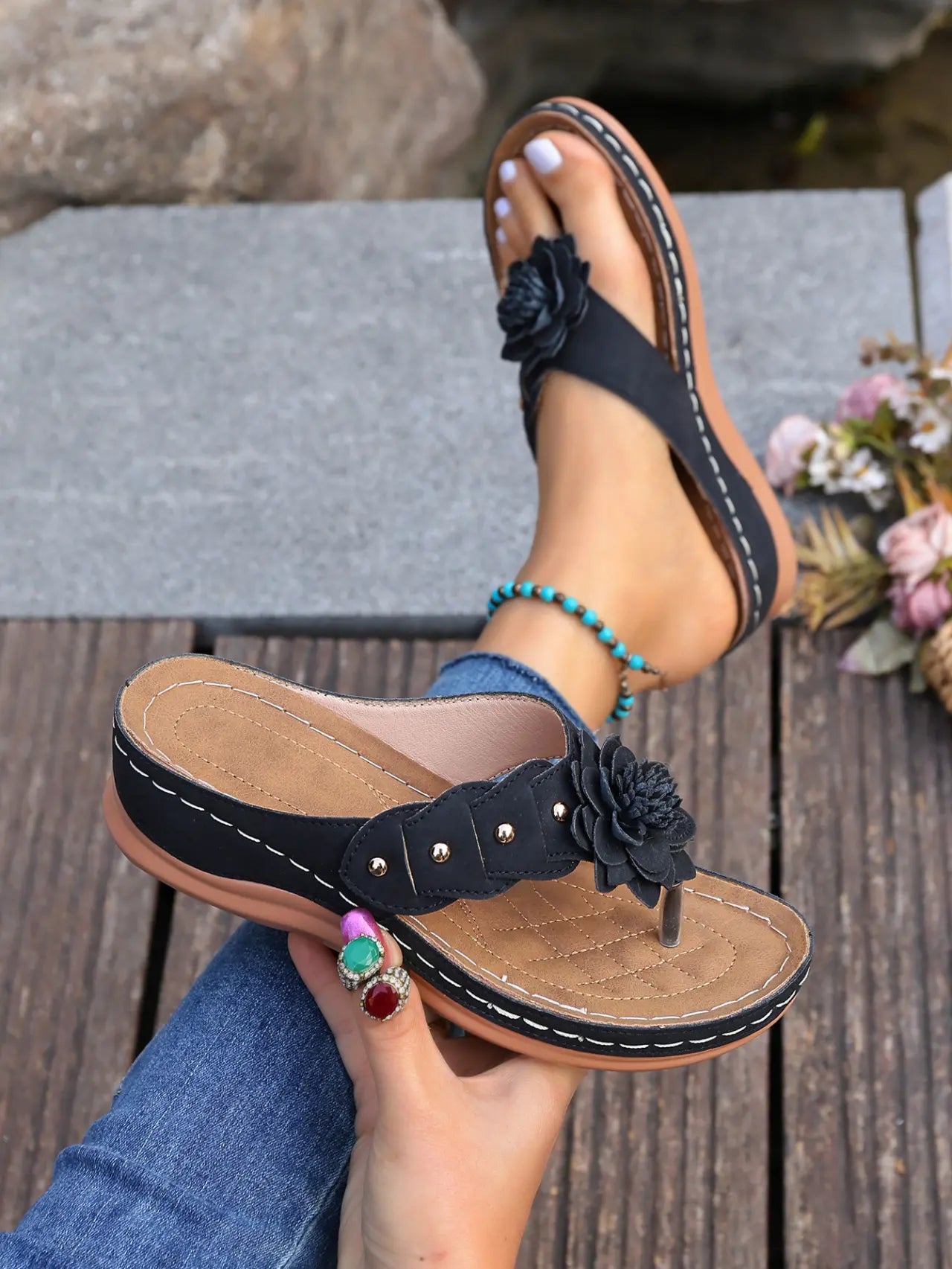 Black Lyraka thong sandals with a black fabric flower embellishment, gold studs, and white contrast stitching, shown on a foot with a turquoise anklet and held in hand, set against a natural outdoor background.