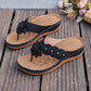 Black Lyraka thong sandals with layered fabric flower embellishments and gold studs, featuring white contrast stitching and a low wedge heel on a textured footbed. Shown on a rustic wooden surface.
